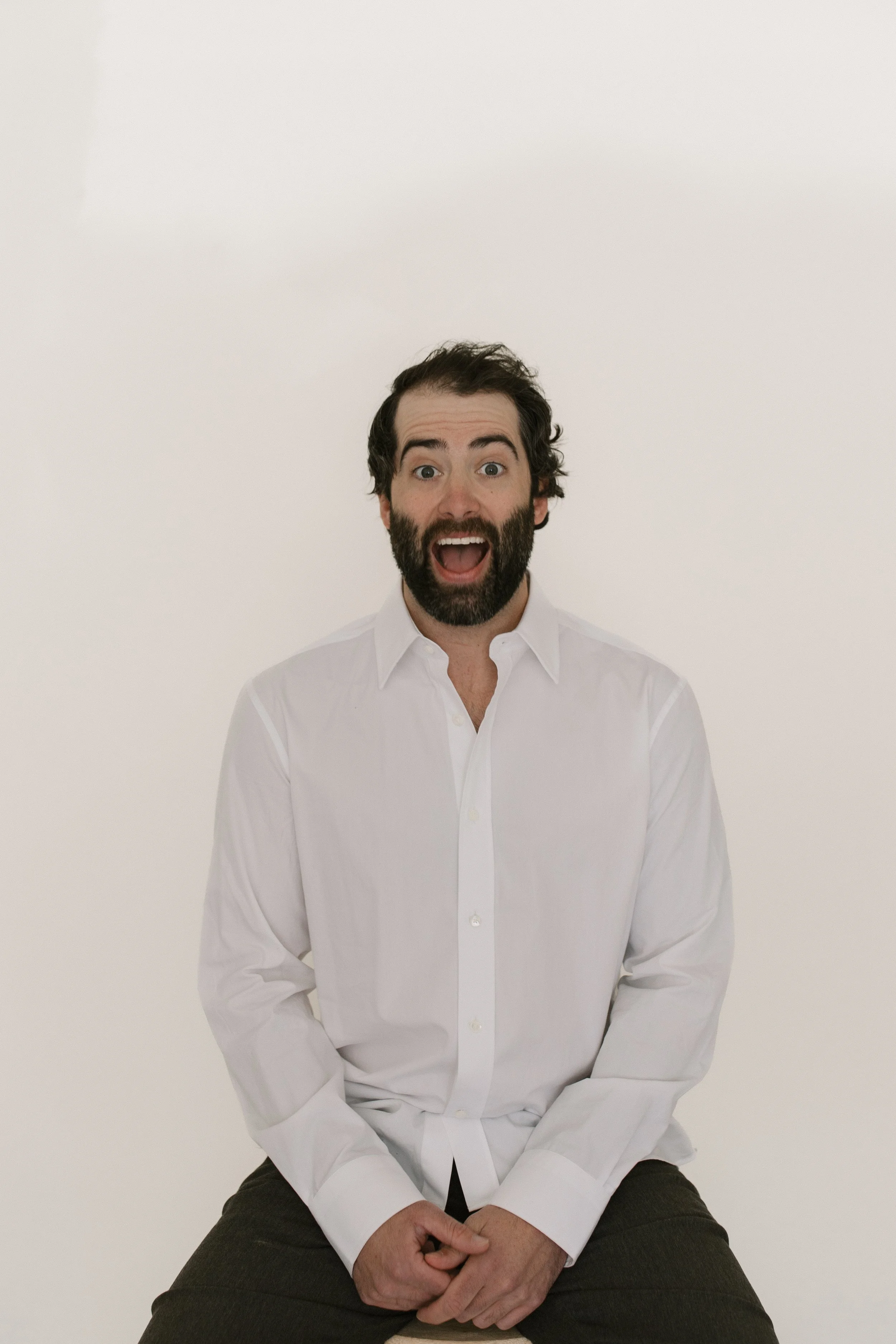 A man with dark hair and beard, wearing a white shirt and sitting against a plain white background, with his mouth open in an expression of surprise or excitement.