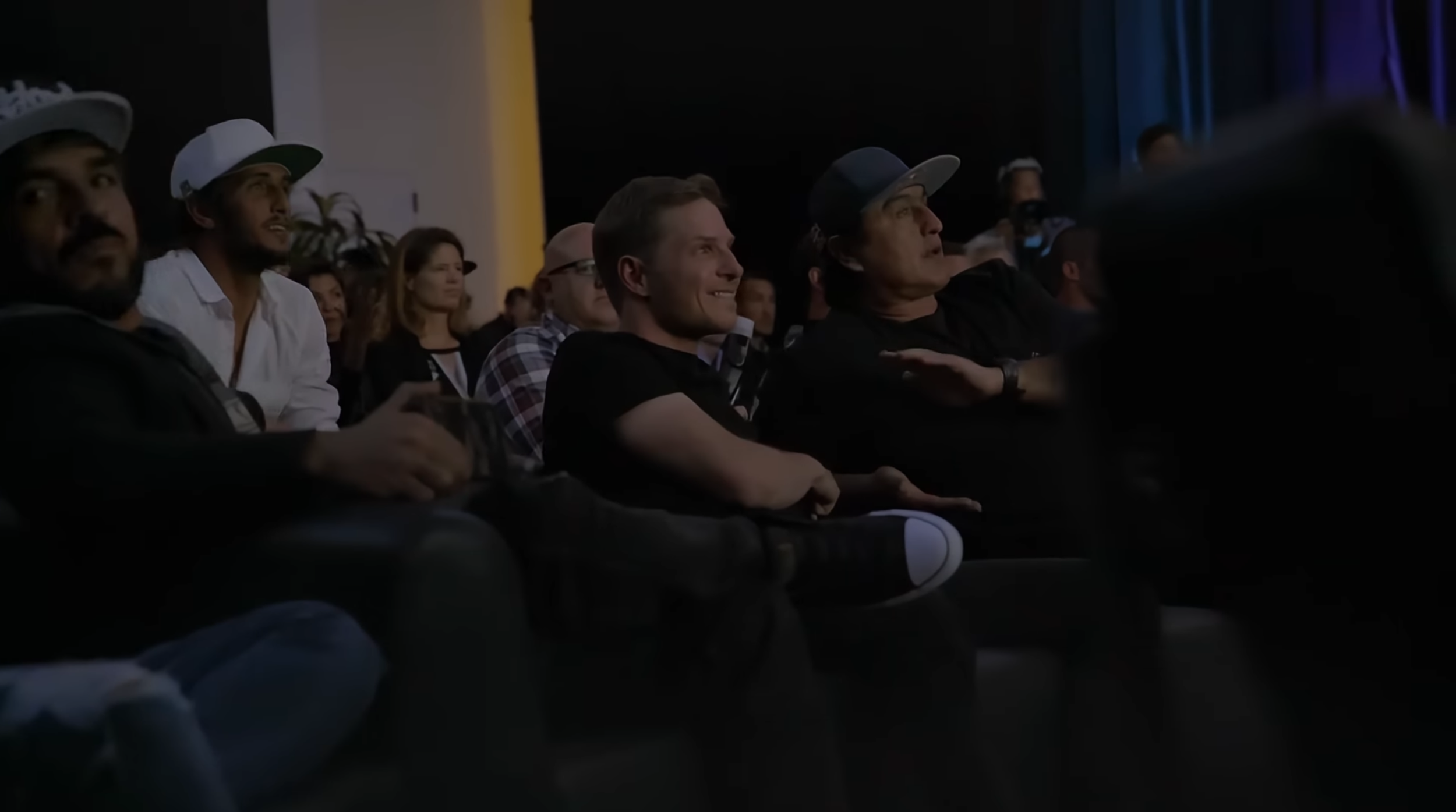 Menschen sitzen in einem dunklen Saal bei einer Veranstaltung, einige lachen und schauen aufmerksam.