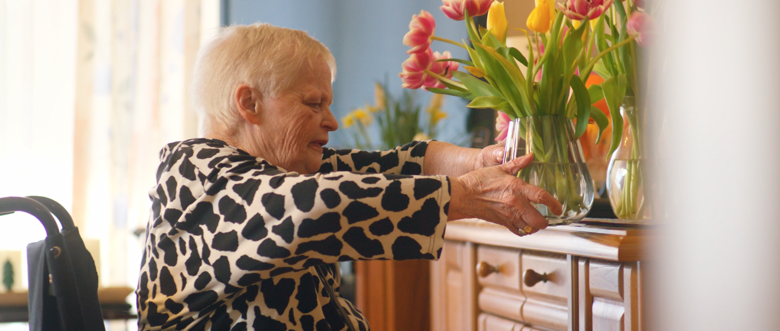 Eine ältere Frau mit kurz blondem Haar, die im Rollstuhl sitzt und einen Blumenstrauß in einer Vase arrangiert, vor einem Holzsideboard, im Hintergrund vor einem Fenster mit codefarbenen Vorhängen.