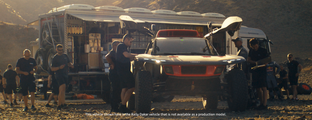 Menschen vor einem Rallye-Fahrzeug und einem großen LKW in einer Wüstenlandschaft, Sonnenlicht, im Hintergrund Berge.