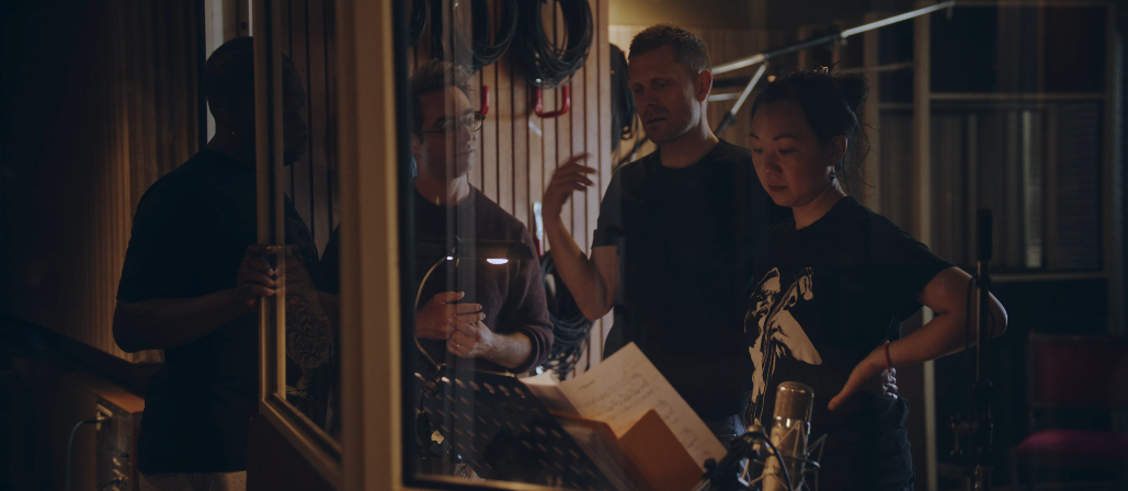 Vier Menschen in einem Tonstudio, die auf die Aufnahme des Musikstücks warten oder es besprechen.