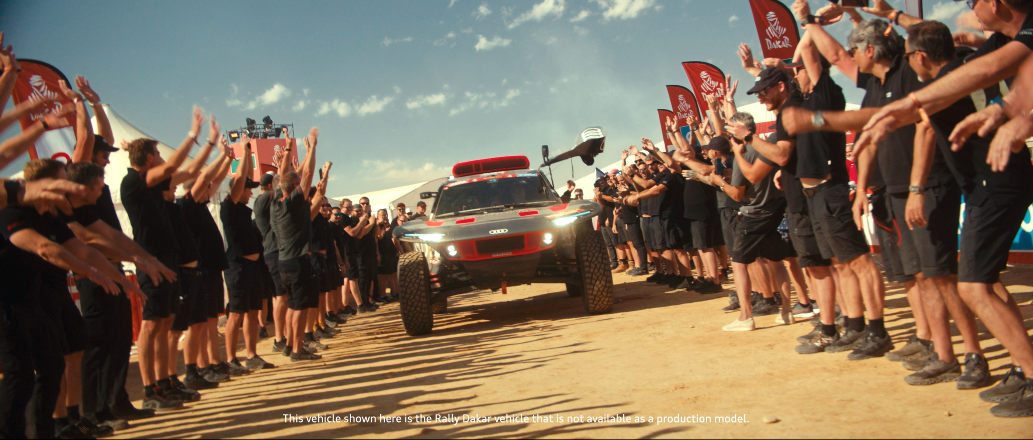 Das Bild zeigt ein Rallye-Auto, das zwischen jubelnden Menschen auf einem Festivalgelände fährt, mit konzentrierter Blickrichtung nach vorne.