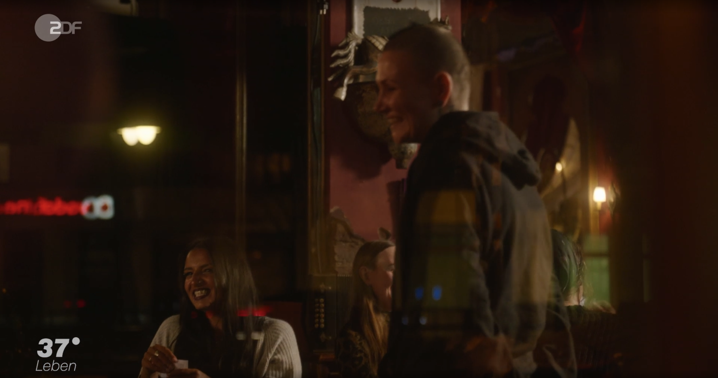 Menschen, die in einem warm beleuchteten Raum lachen und sich unterhalten, Blick durch eine Fensterscheibe