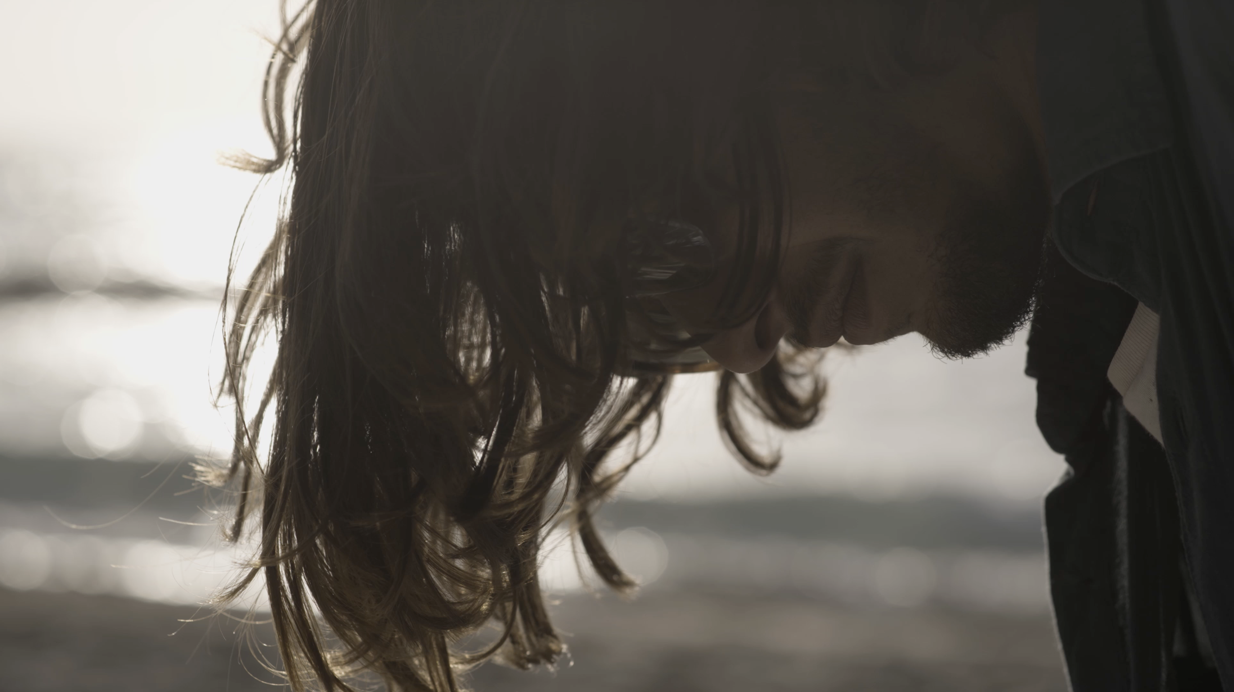 Nahaufnahme eines Mannes mit langen, lockigen Haaren und Brille, der nach unten schaut. Im Hintergrund sind unscharfe Lichter.