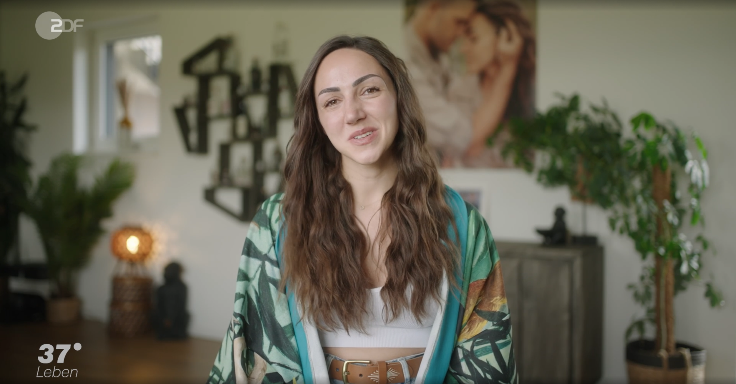 Junge Frau mit lang, lockigem braunem Haar, die vor einer Kamera sitzt in einem hellen Wohnzimmer mit Pflanzen und Kunstwerken im Hintergrund.