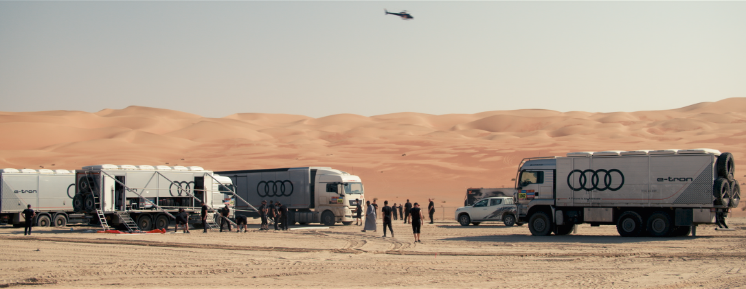 Menschen und Fahrzeuge in der Wüste, mit mehreren großen LKWs, einer Bohrmaschine und einem Helicopter am Himmel.