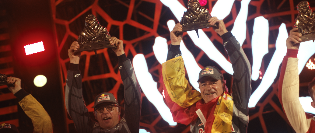 Menschen feiern auf einem Podium, die den Sieg bei einem Motorsport-Event feiern, einige tragen Red Bull Kappen.