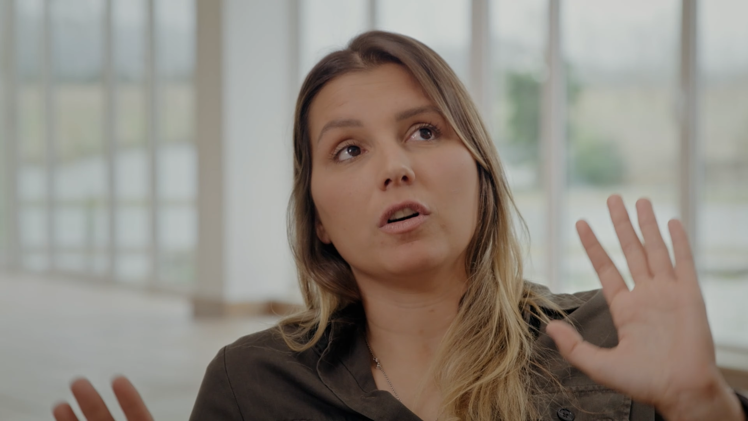 Eine Frau wirkt, als ob sie etwas erklärt oder erklärt wird, in einem hellen Raum mit großen Fenstern im Hintergrund.