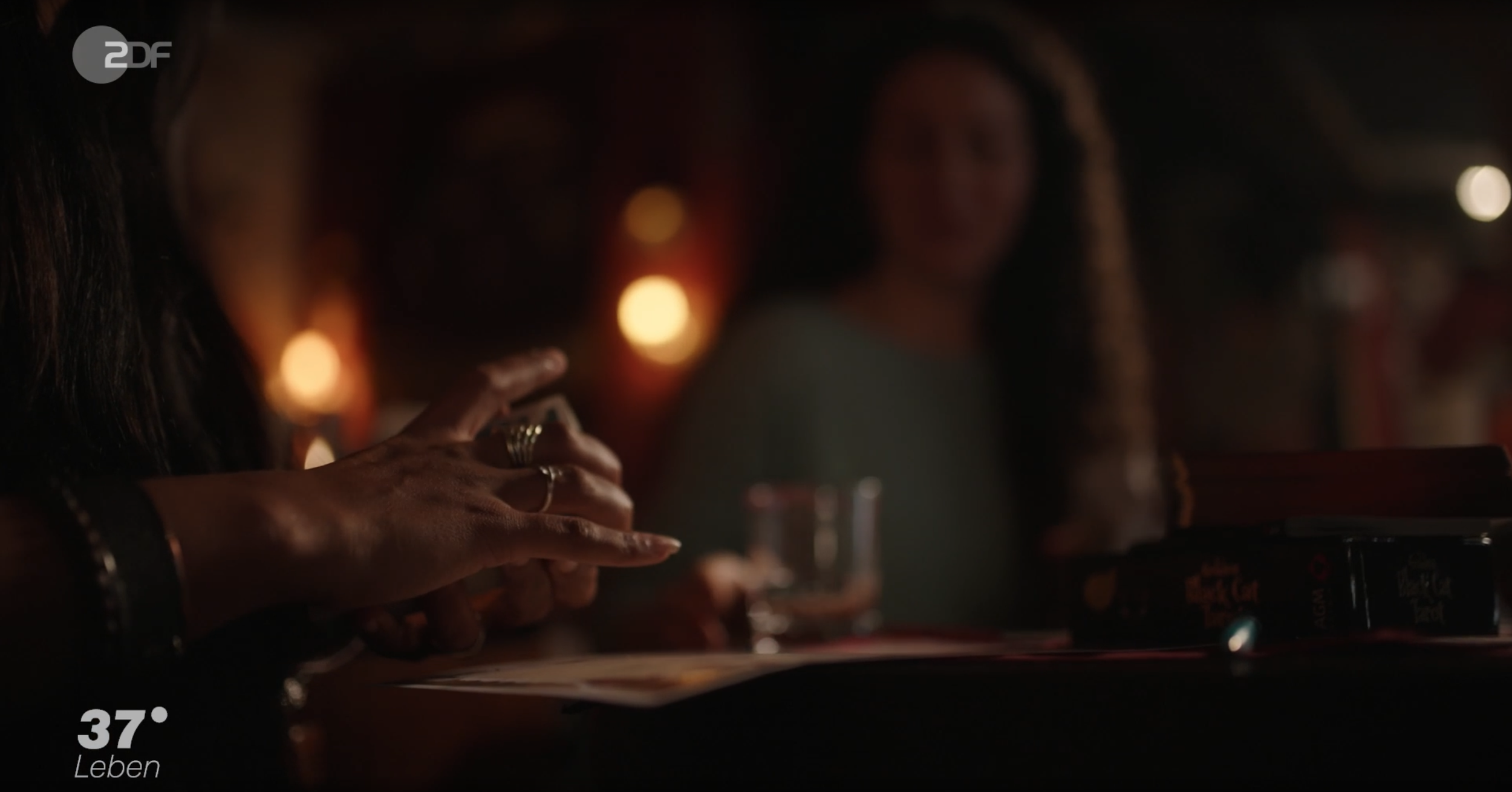 Nahaufnahme einer Hand mit mehreren Ringen, die auf einem Tisch liegt, im Hintergrund eine lächelnde Frau in einem düsteren Ambiente mit warmem Licht.