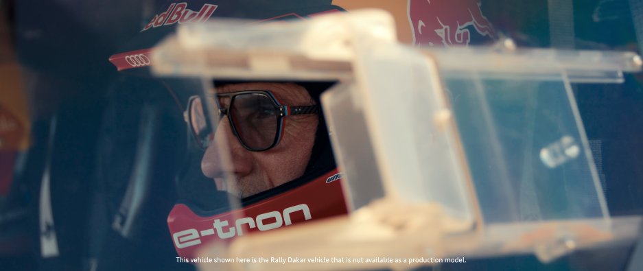 Nahaufnahme eines Mannes in einem Rennfahrzeug, sichtbar durch das Visier des Helms, mit Red Bull und Rally Dakar Branding.