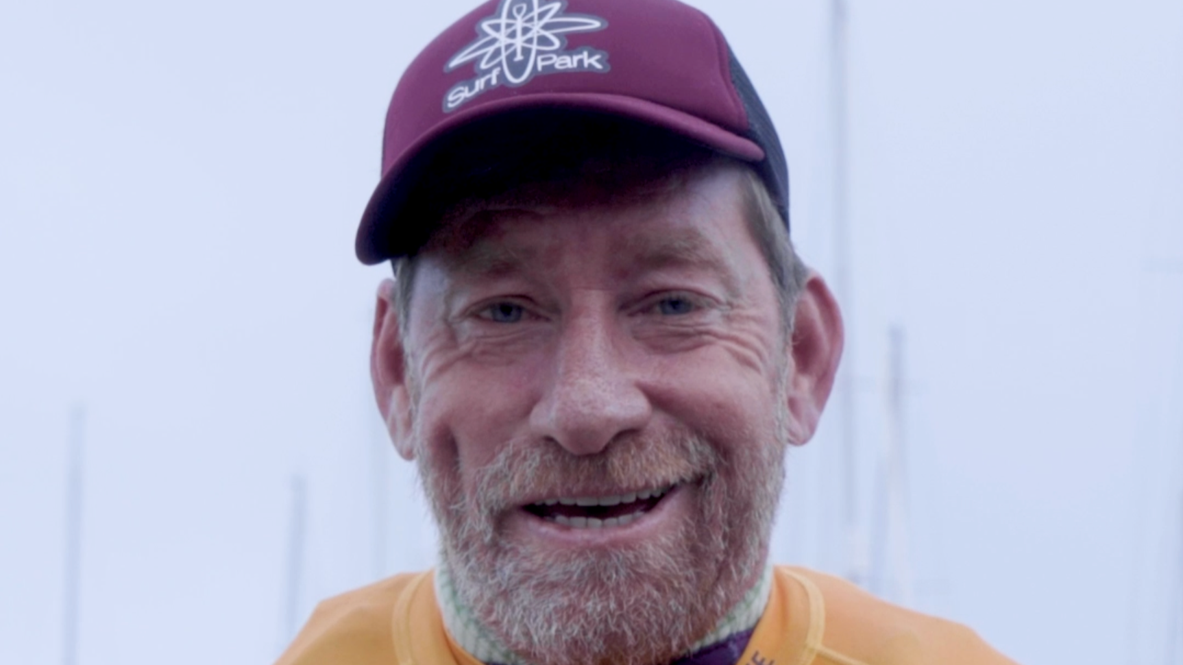Ein lachender Mann mit einem roten Schnurrbart und grauem Bart, trägt eine rote Kappe mit einem Surfdampfer-Logo, trägt ein gelbes Shirt, im Hintergrund sind Masten eines Segelbootes vor einem nebligen Himmel.