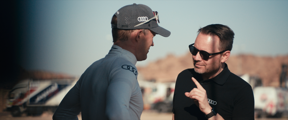 Zwei Männer im Gespräch, einem trägt einen Rennfahrerhelm, der andere trägt Sonnenbrille, im Hintergrund eine Wüstenlandschaft und Fahrzeuge.