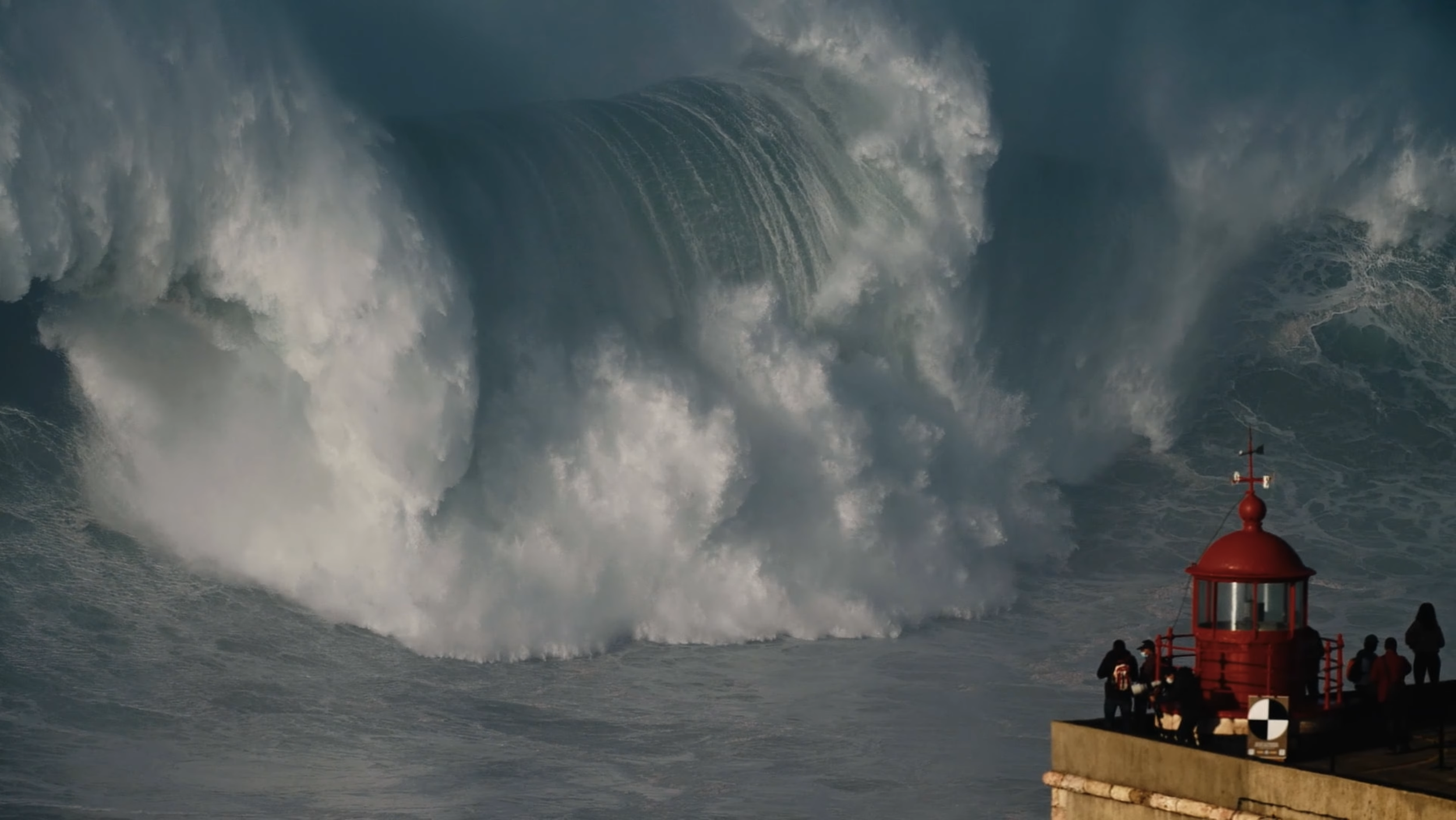 Große Wellen schlagen gegen einen Leuchtturm an einer Küste, mit mehreren Personen auf dem Dach des Leuchtturms