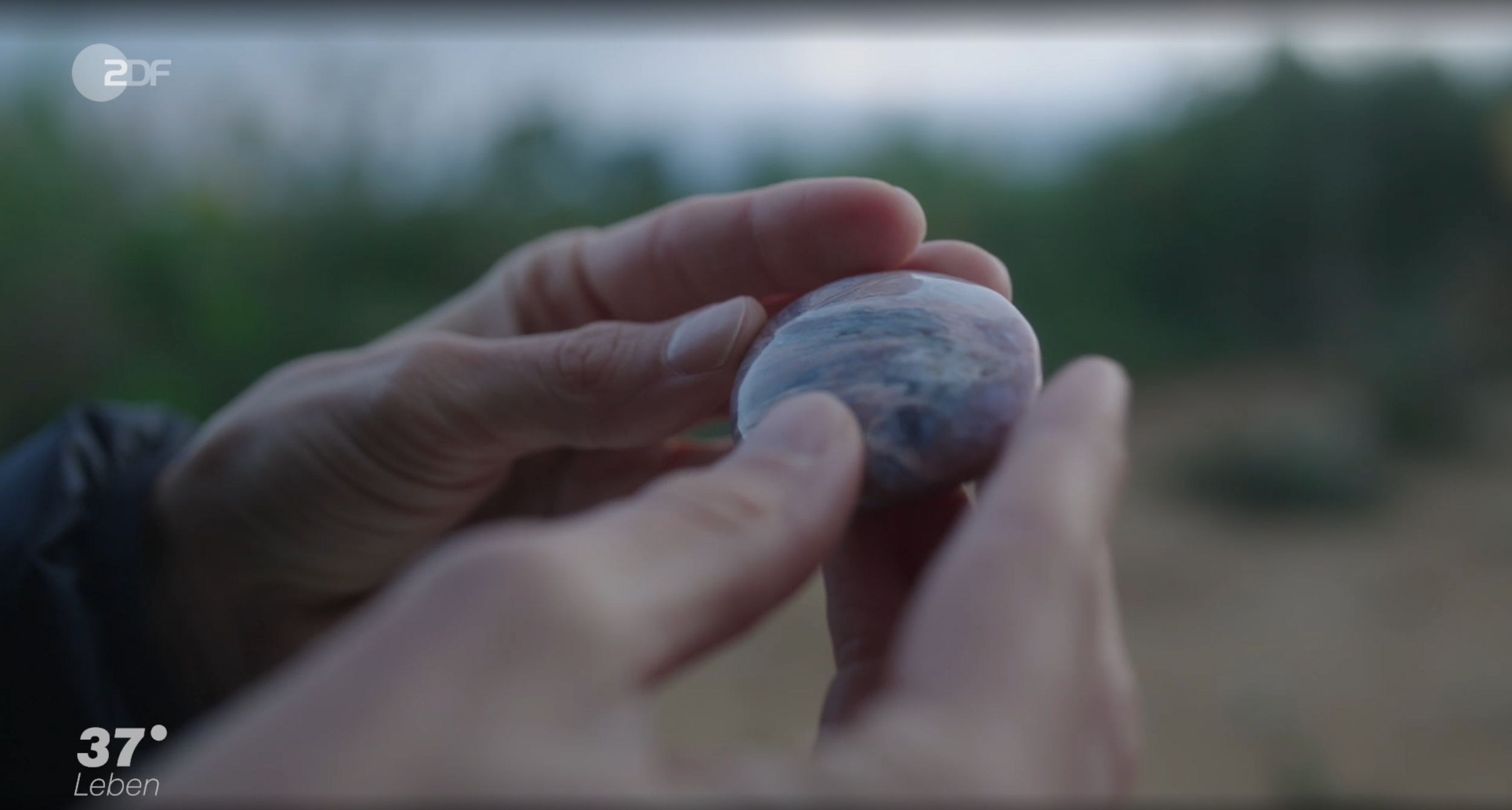 Nahaufnahme einer Hand, die einen Edelstein hält, im Hintergrund unscharfe Natur. Der obere linke Bereich zeigt das Logo von ZDF.