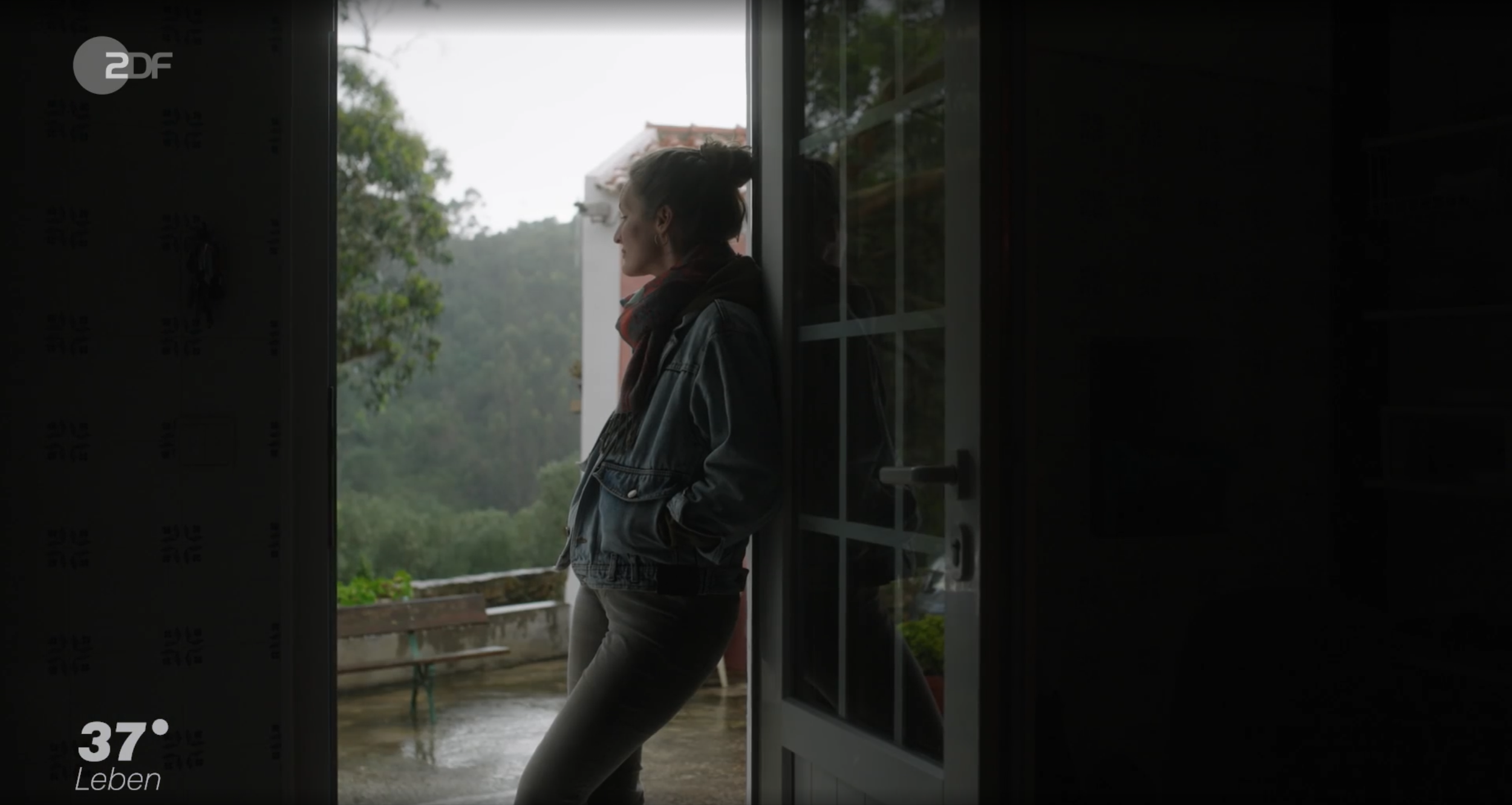 Frau steht im Türrahmen und blickt nach draußen bei Regen, auf einem Balkon im Grünen.