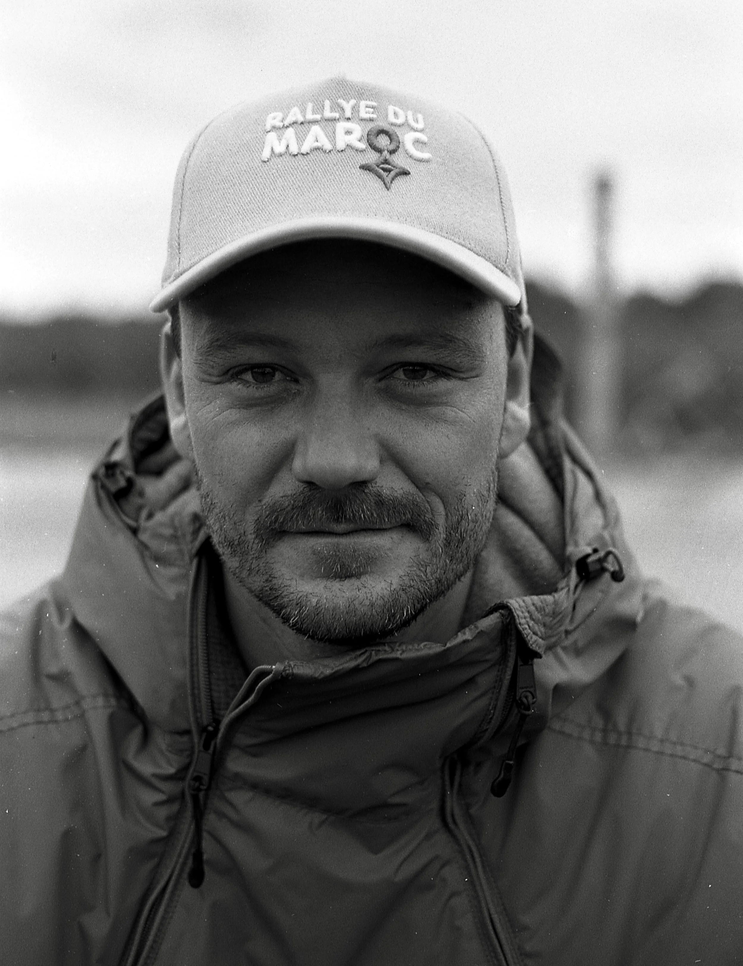Ein Mann trägt eine Mütze mit dem Schriftzug 'Rallye du Maroc' und eine wetterfeste Jacke, Blick direkt in die Kamera, im Hintergrund unscharf Natur.