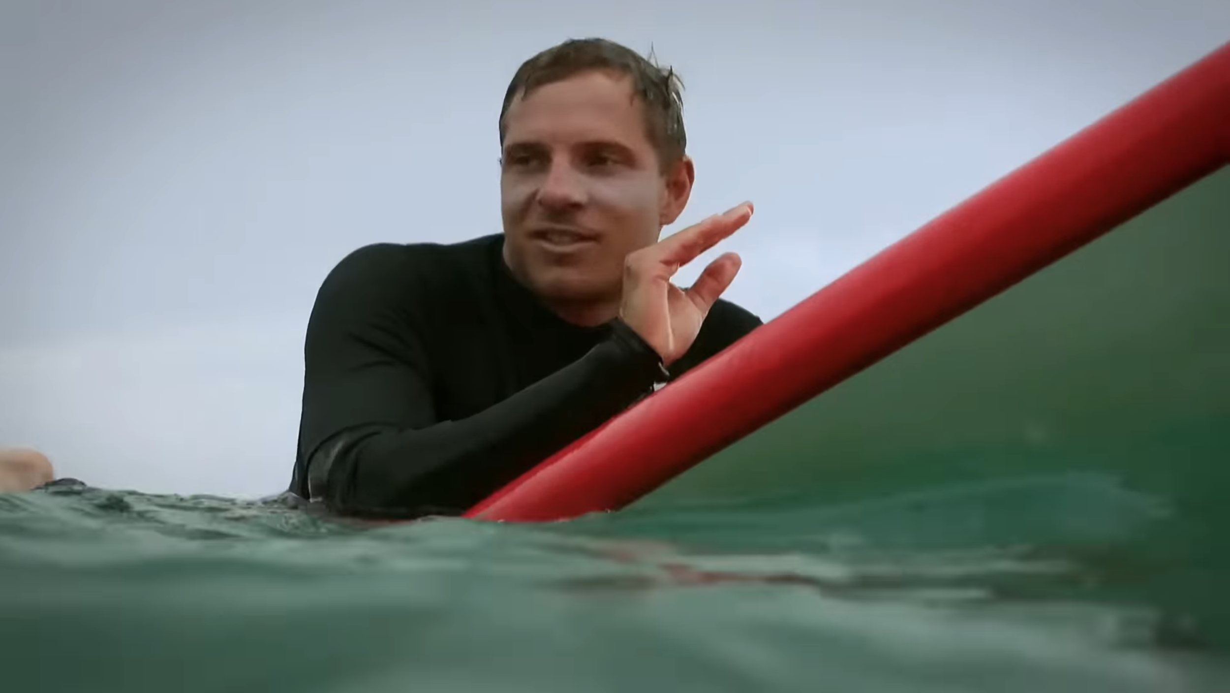 Ein Mann im schwarzen Neoprenanzug sitzt auf einem Surfbrett im Wasser und spricht. Der Himmel ist bewölkt.