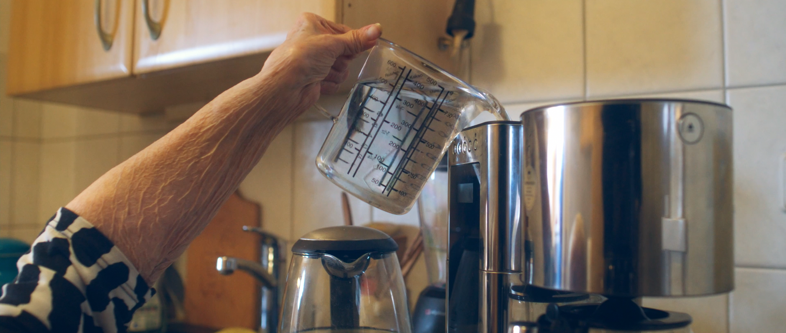 Eine ältere Person gießt Wasser aus einem Messbecher in eine Kaffeemaschine in einer Küche.