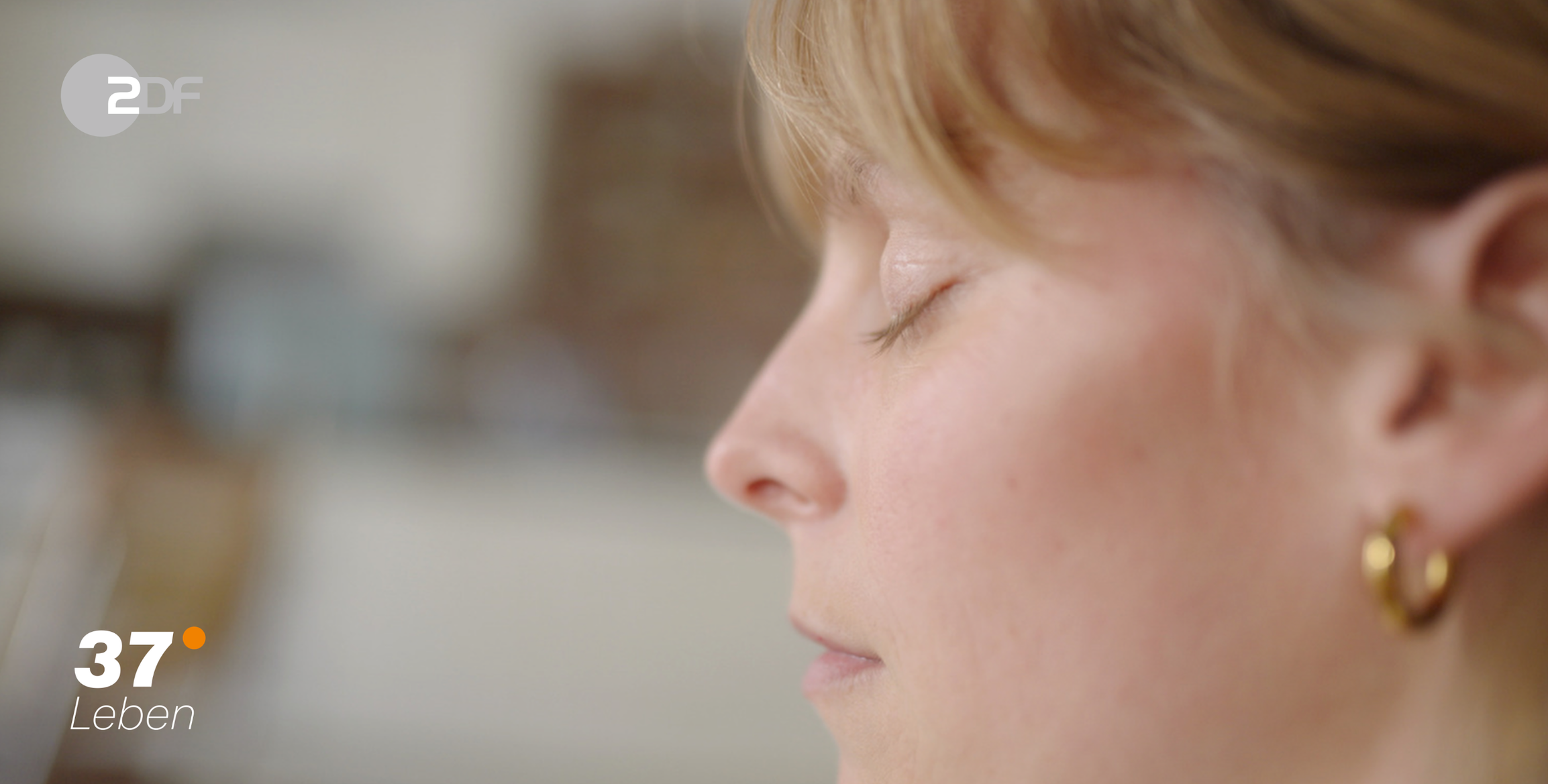 Nahaufnahme eines jungen Mädchens mit geschlossenem Blick, kurze blonde Haare, Goldohrringe, entspannter Gesichtsausdruck.