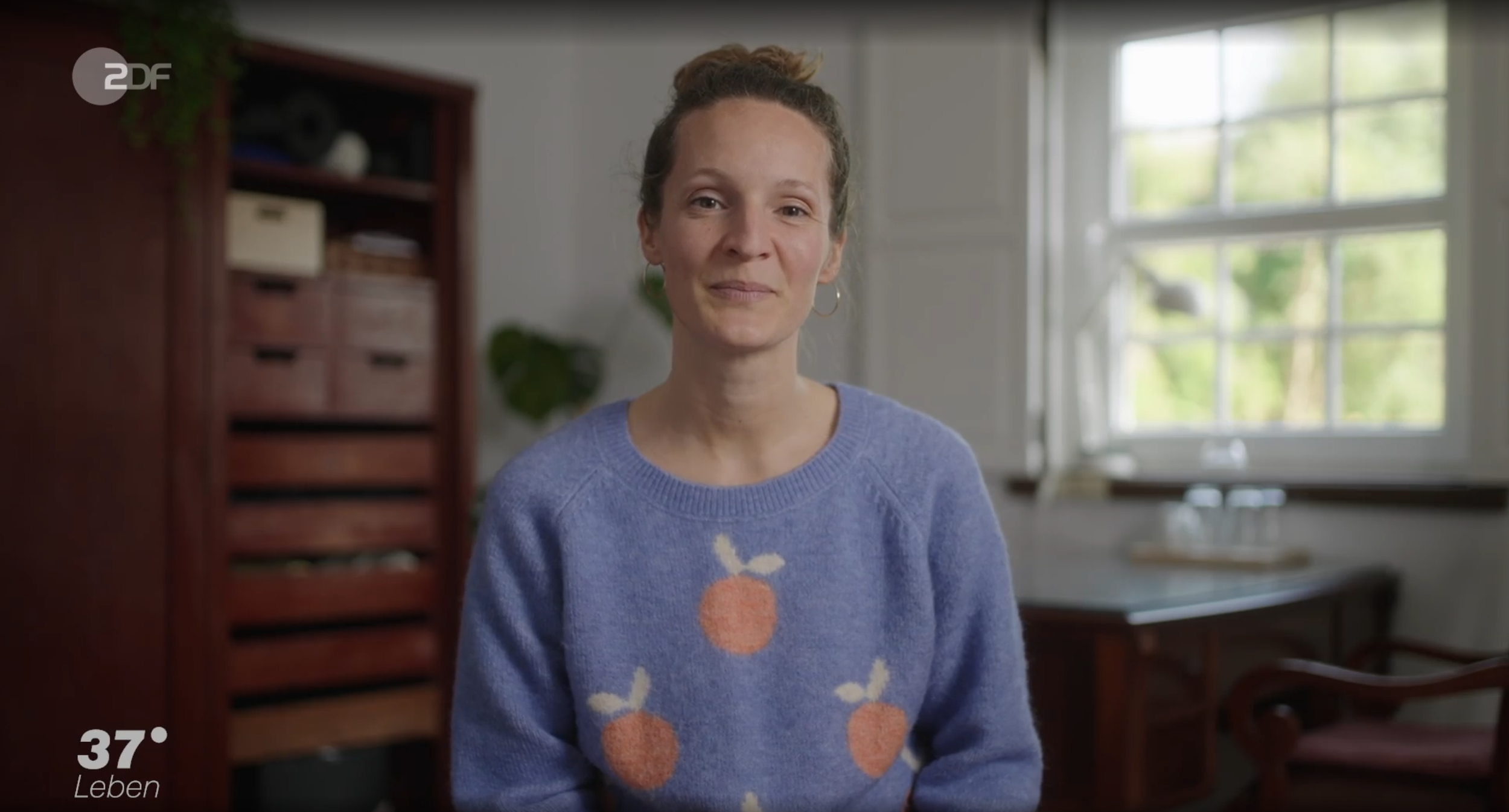 Eine Frau sitzt in einem hellen Raum mit großen Fenstern, trägt einen blauen Pullover mit Apfelmotiven, im Hintergrund sind Holzregale und eine Tisch mit einem Wasserkrug und Gläsern.