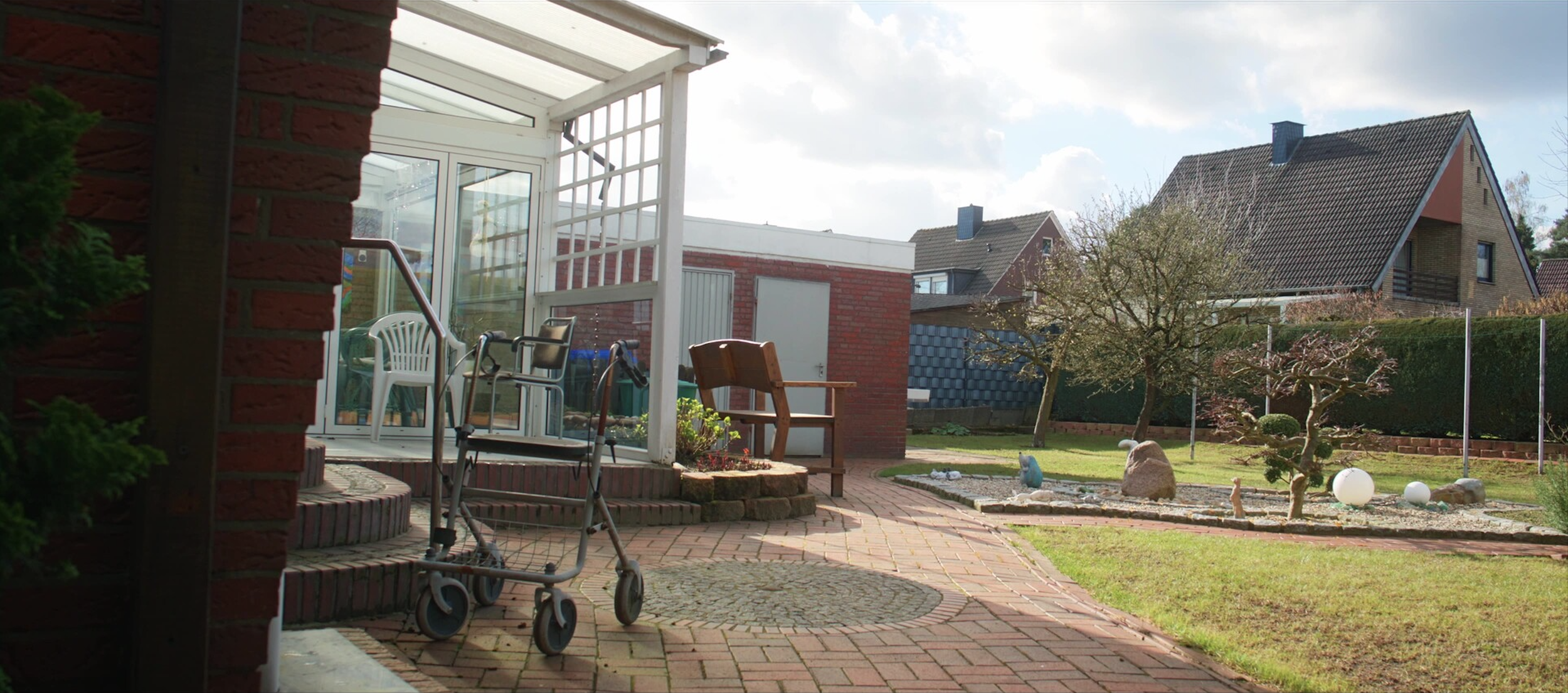 Ein gepflegter Garten mit Terrassenbereich, neleinem Baum, Steine und Kugeln als Gartendekoration, umgeben von Häusern mit Ziegeldächern.
