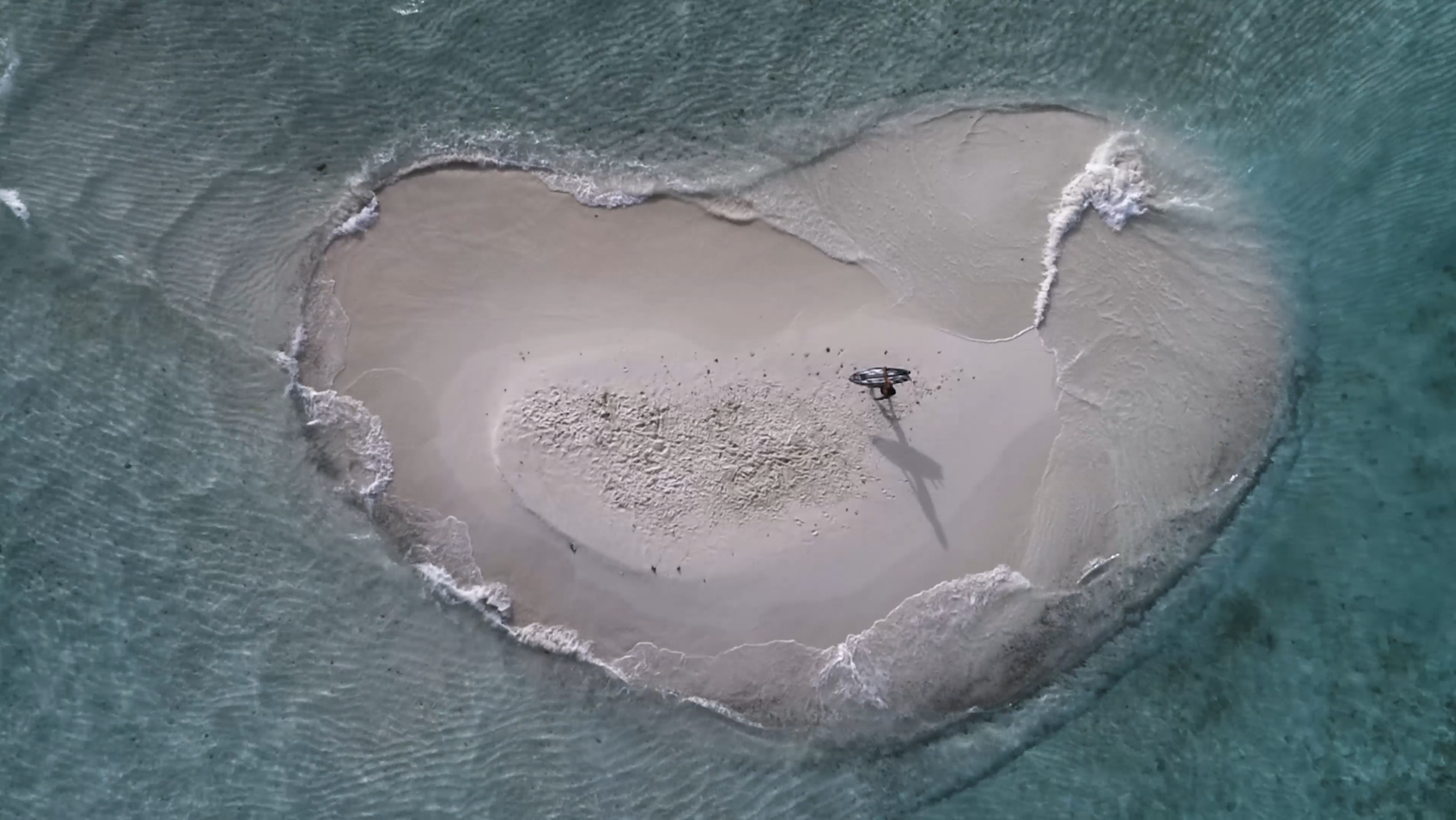 Vogelperspektiveaufnahme einer kleinen, sandigen Insel inmitten des Meeres mit einem Surfer, der auf der Insel steht.
