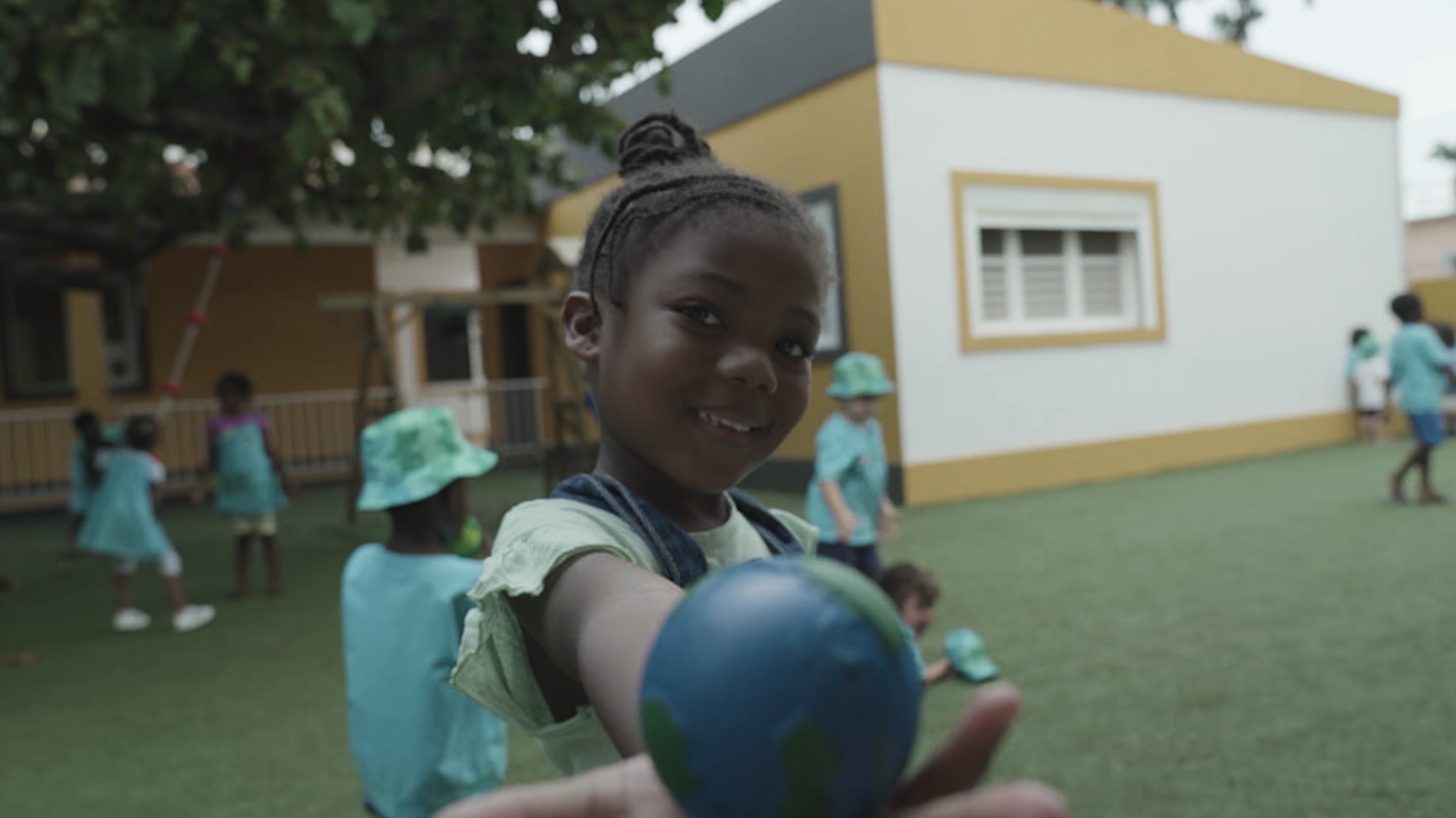 Ein lachendes Mädchen, das auf einem Spielplatz eine kleine Weltkugel hält, im Hintergrund andere Kinder und ein Haus.