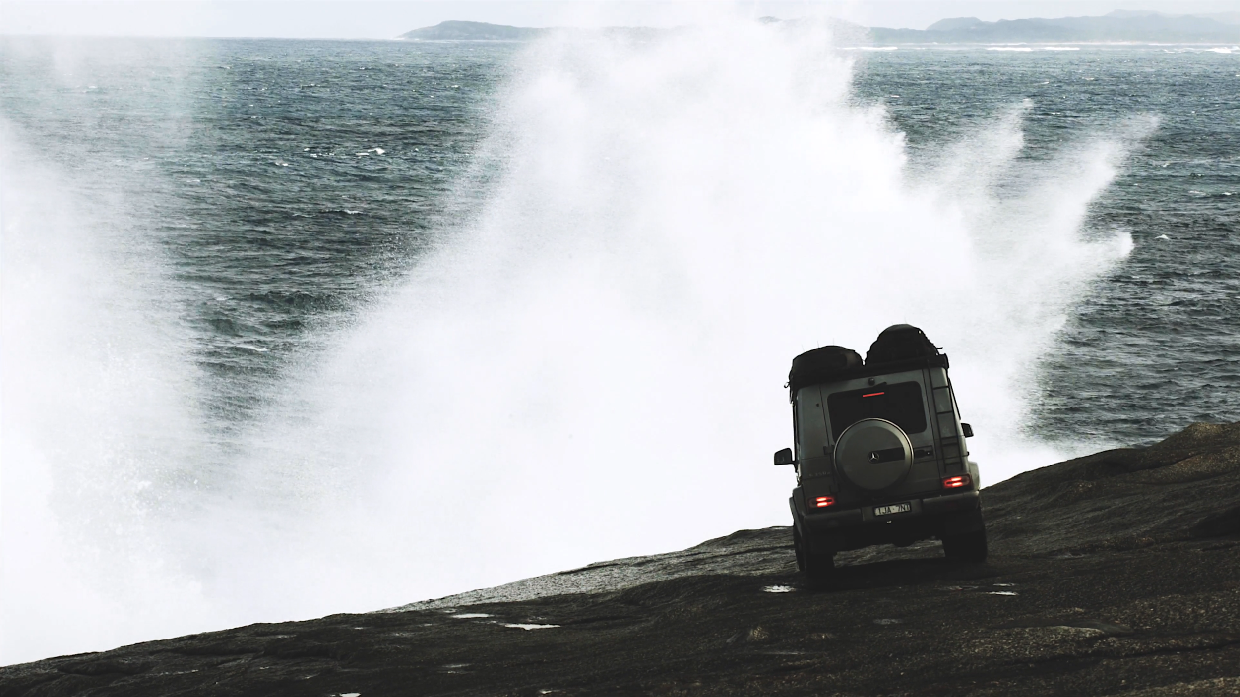Ein schwarzer Geländewagen steht auf einer felsigen Küstenlinie, während hohe Wasserwellen gegen die Felsen schlagen.