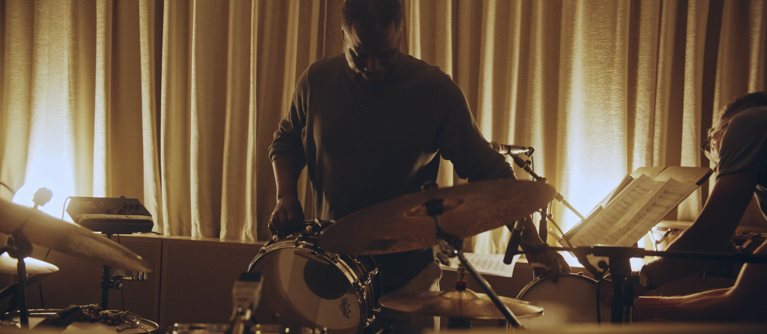 Musiker spielt Schlagzeug in einem Raum mit gelben Vorhängen im Hintergrund.