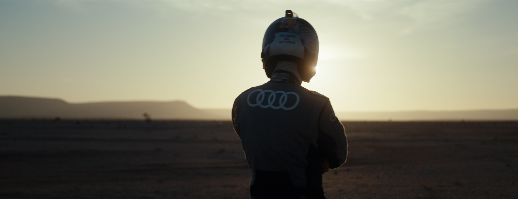 Ein Pilot mit einem schwarzen Helm steht im Wüstensand bei Sonnenuntergang, mit Audi-Logos auf der Kleidung.