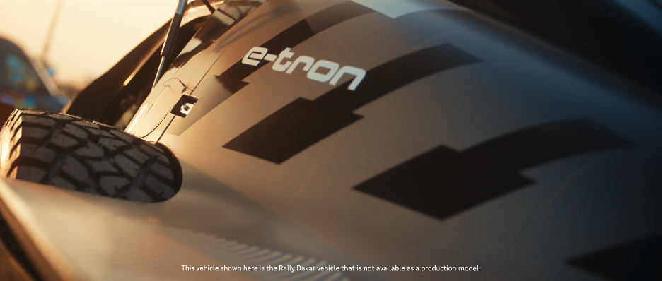 Nahaufnahme eines Rallye-Fahrzeugs mit dem Logo 'e-tron' auf der Seite, mit einem Reifen im Vordergrund, im Sonnenlicht aufgenommen.