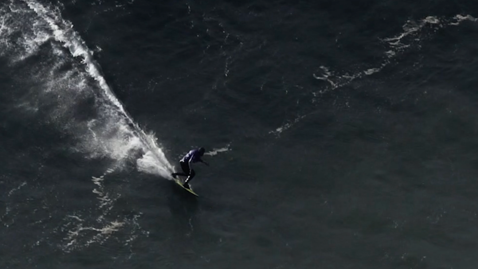 Surfer auf einem schwarzen Surfbrett am Meer bei hohem Wellengang, Wasser spritzt um ihn herum.