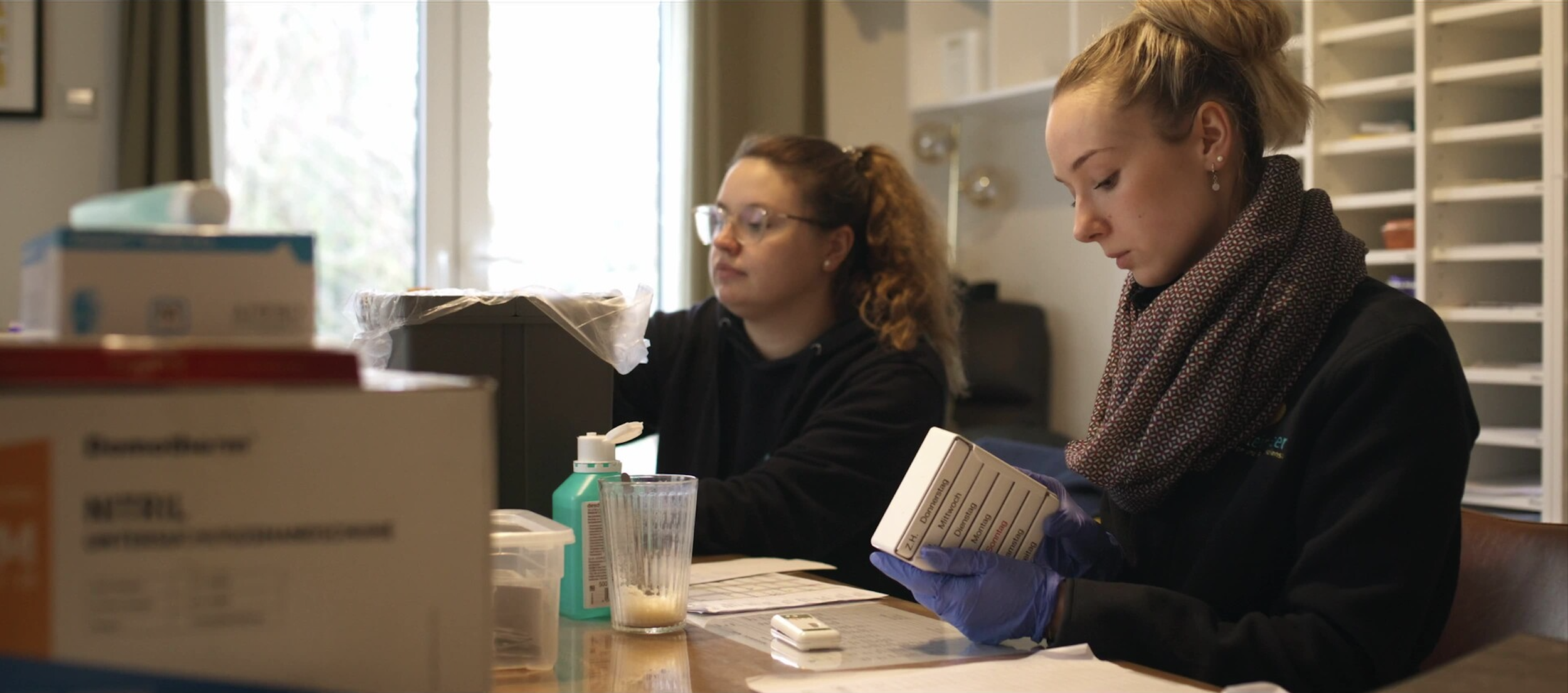 Zwei Frauen arbeiten an einem medizinischen Projekt in einem Raum mit Regalen im Hintergrund. Sie verwenden Medikamente und medizinische Utensilien auf dem Tisch.