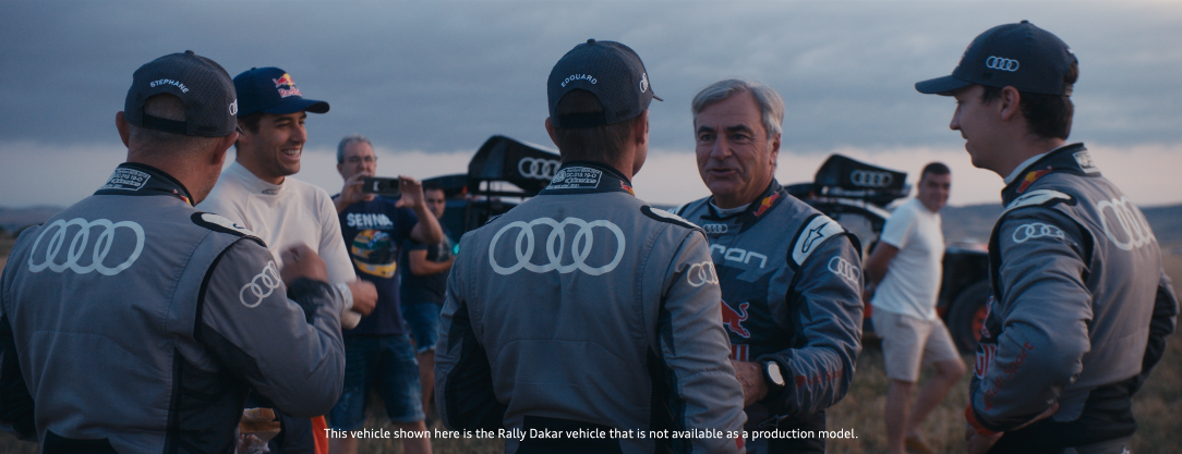 Motorsport-Teammitglieder und Fahrer im Gespräch auf einer Rennstrecke, mit Rallye-Auto im Hintergrund, bei Dämmerlicht.