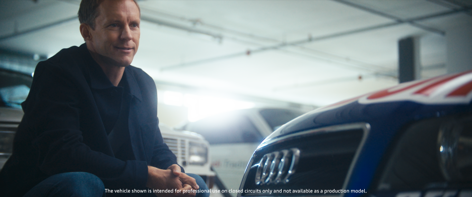 Ein Mann sitzt in einer Tiefgarage zwischen Autos, darunter ein Audi. Er trägt dunkle Kleidung und schaut nachdenklich in die Ferne.