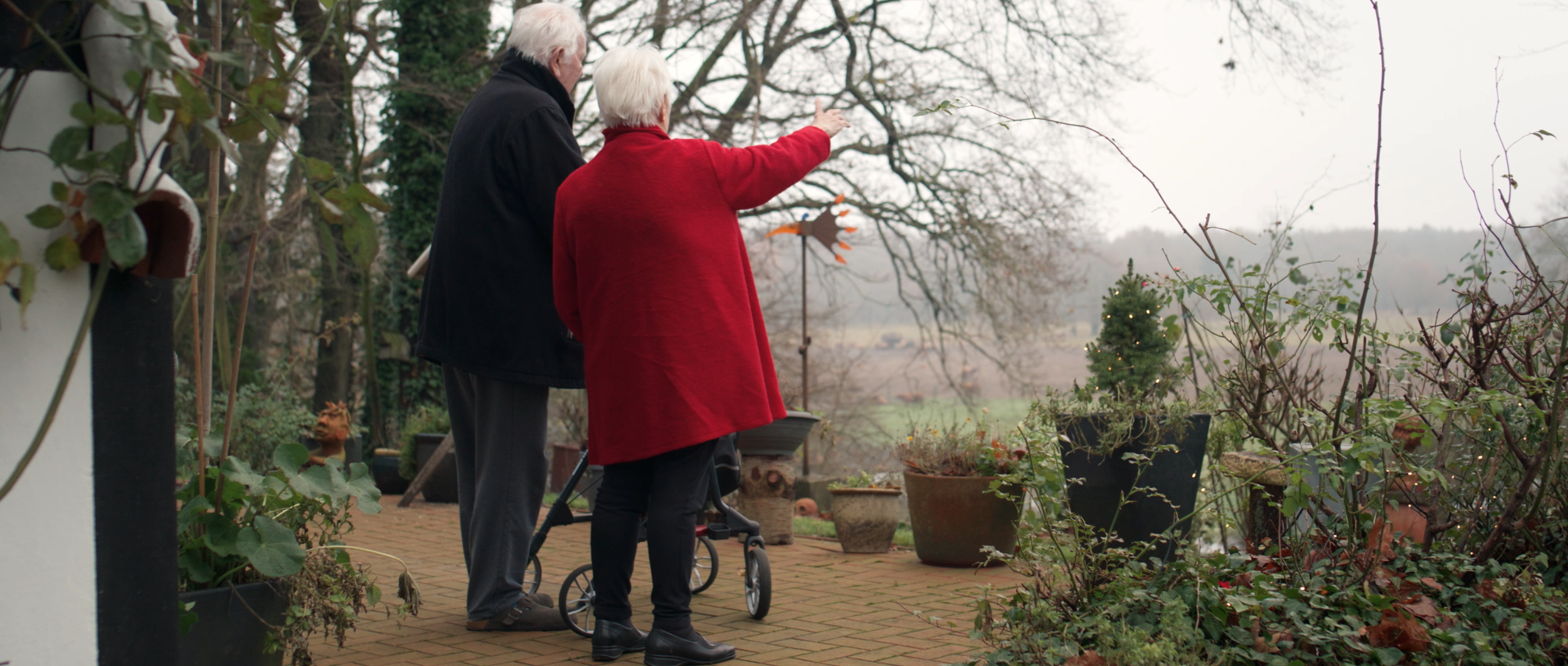 Älteres Paar auf einem Balkon im Gespräch, mit einem Rollator, umgeben von Pflanzen und Töpfen, Blick in die ferne Landschaft bei bewölktem Himmel.