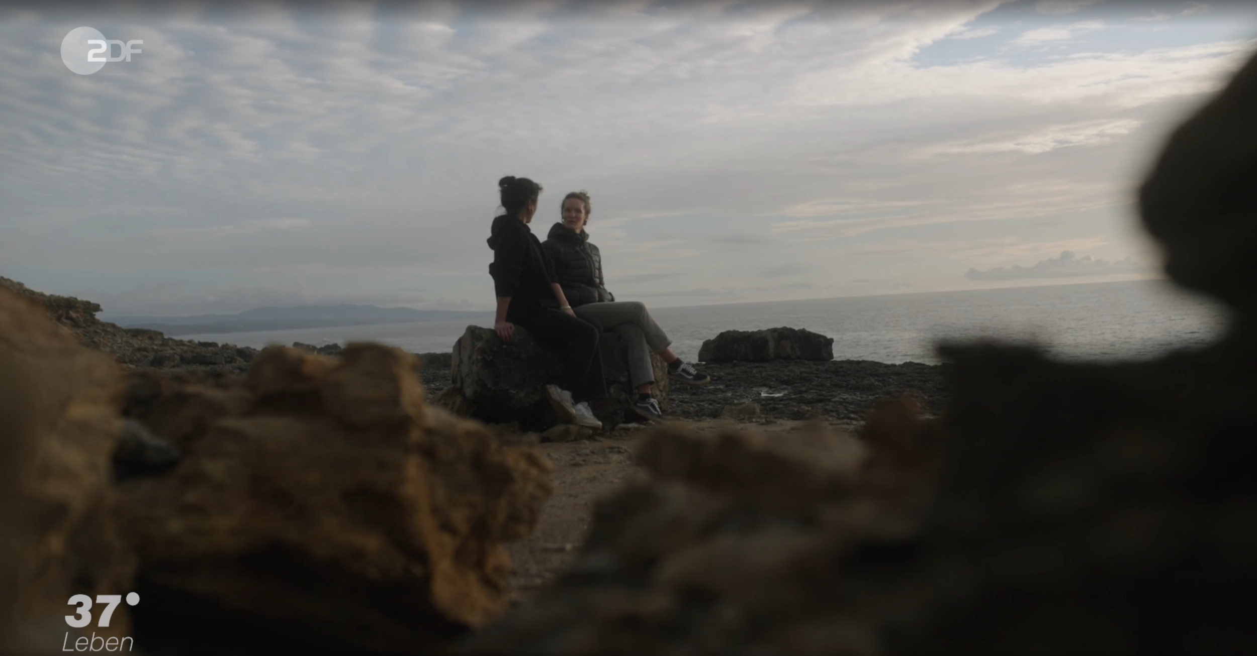 Zwei Frauen sitzen auf einem Felsen am Meer und unterhalten sich, während die Kamera sie aus einer niedrigen Perspektive durch Steine filmt.