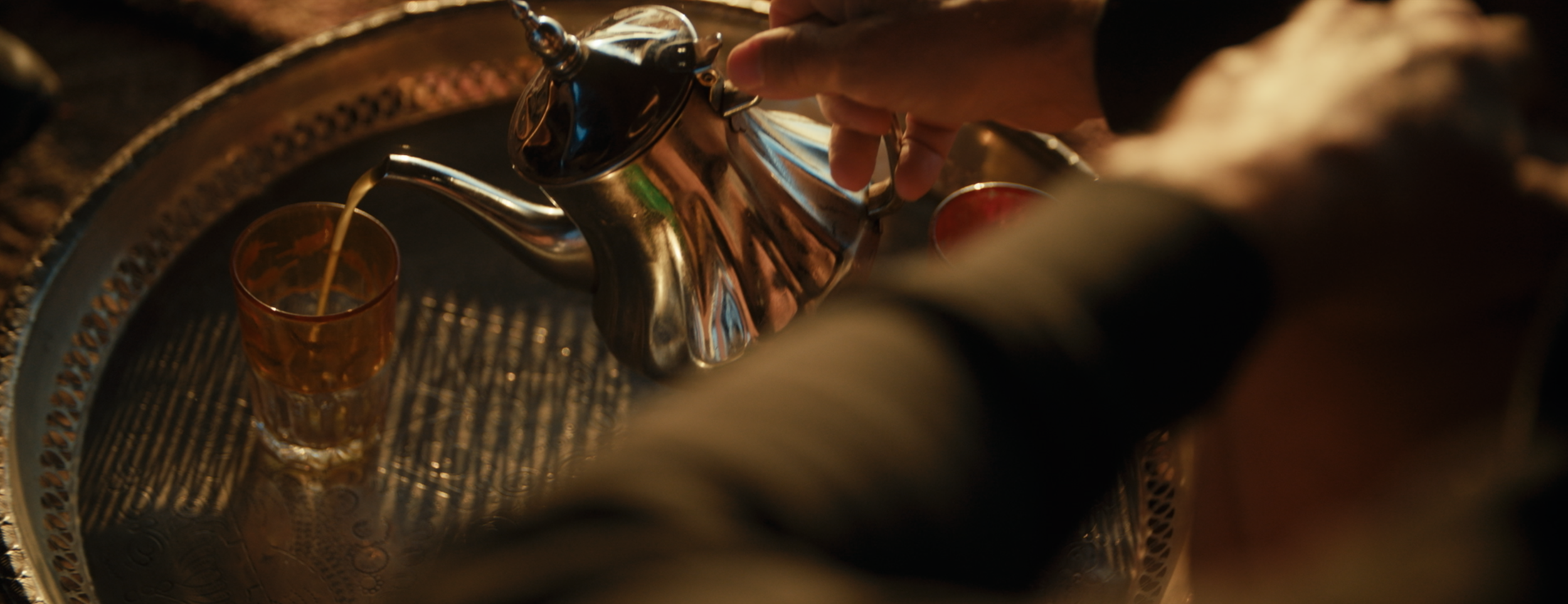 Eine Person gießt Tee aus einer glänzenden Metallkanne in ein Glas. Das Setting ist warm beleuchtet, und das Metalltablett spiegelt das Licht. Der Fokus liegt auf der Hand, die den Teekannen-Hahn hält.