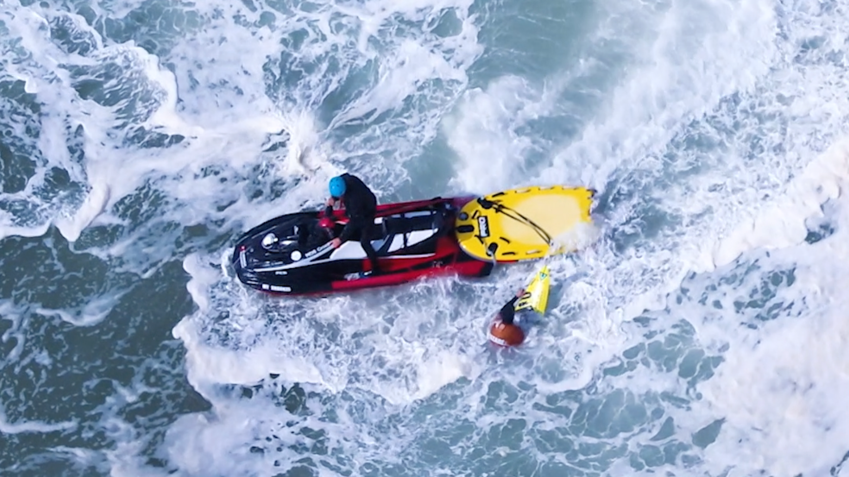 Ein Mensch auf einem Jet-Ski in den Wellen des Meeres, trägt einen blauen Helm und einen schwarzen Anzug, ein anderer Mensch mit einem orangefarbenen Helm hält ein gelbes Surfbrett im Wasser.