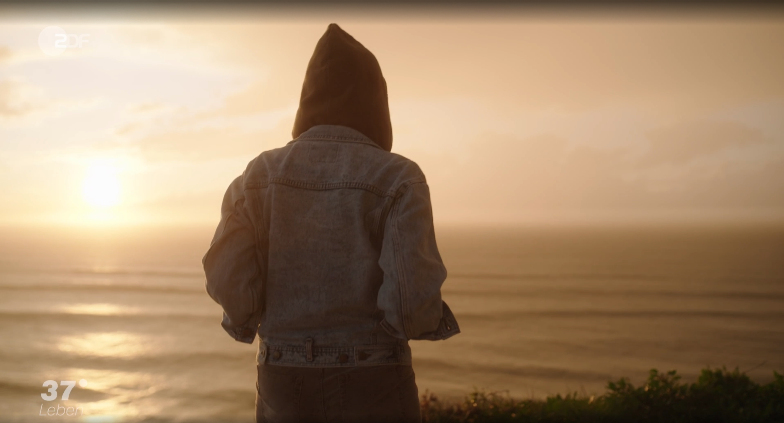 Person mit Kapuze in einer Jeansjacke blickt auf das Meer während eines Sonnenuntergangs.