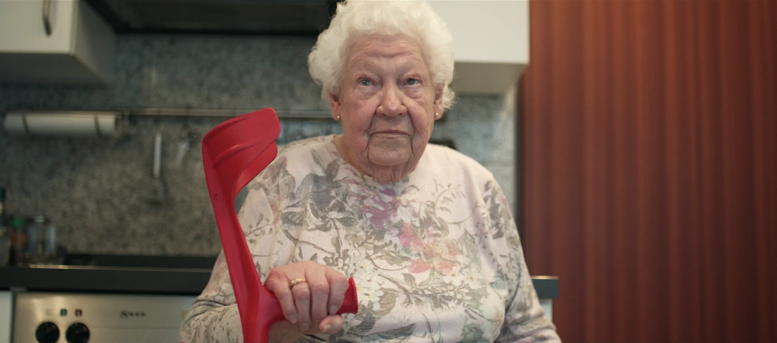 Ältere Frau mit weißen Haaren hält einen roten Gegenstand in der Hand, steht in einer Küche mit dunklem Küchenhaus und Holzvertäfelung.