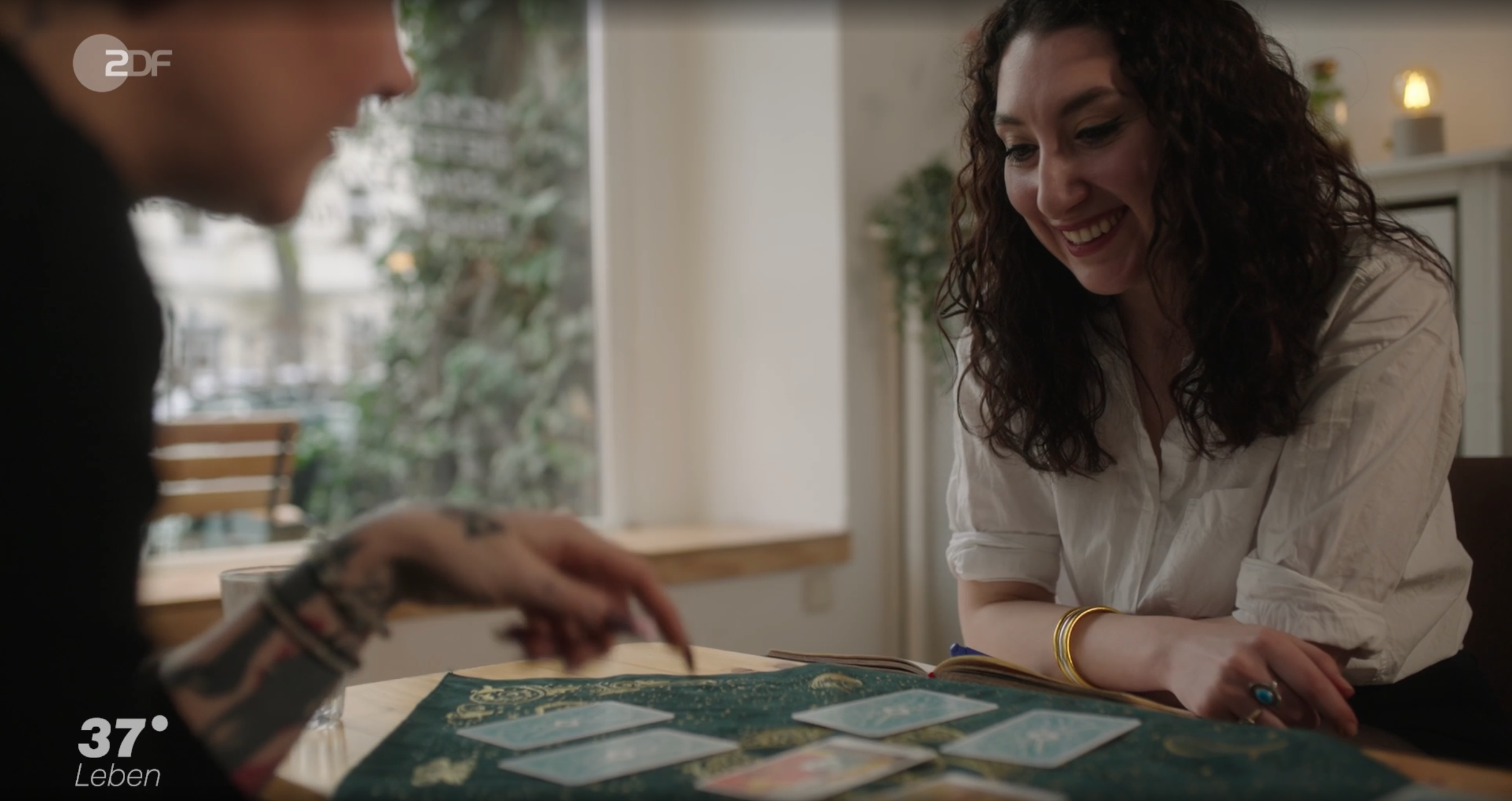 Zwei Frauen spielen Tarotkarten an einem Tisch. Die Frau auf der rechten Seite lächelt, während die andere Frau nur teilweise sichtbar ist.