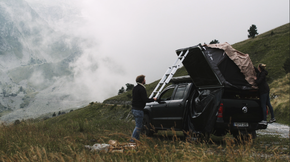Zwei Personen bauen ein Zelt auf einem schwarzen Pickup-Truck in einer Berglandschaft auf, umgeben von Gras und Bäumen.