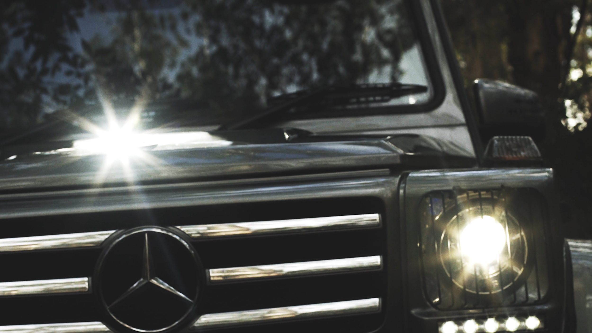 Nahaufnahme der Front eines schwarzen Mercedes-Benz Geländewagens mit leuchtendem Scheinwerfer, im Hintergrund Bäume und Himmel