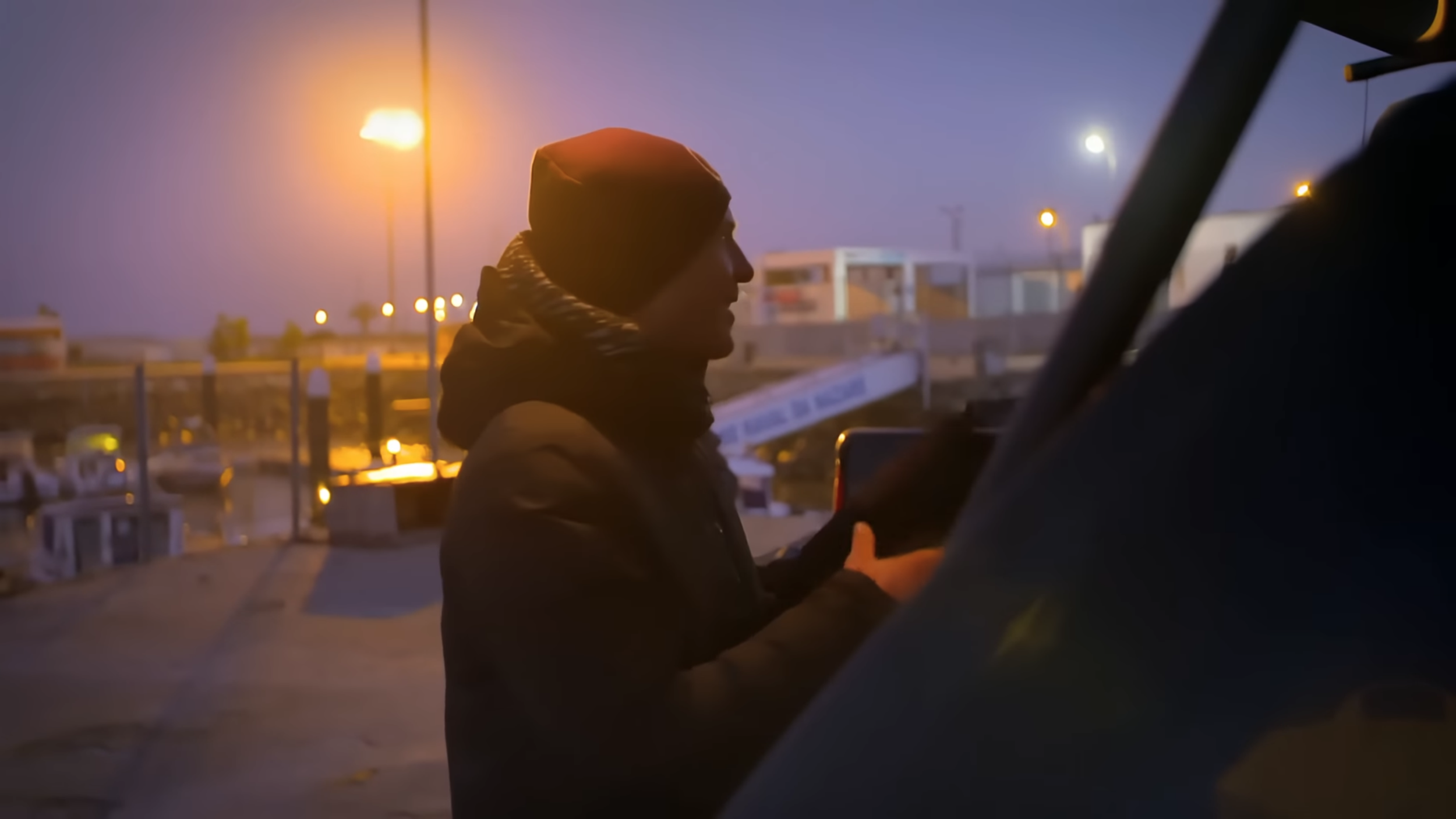 Junge Person in dunkler Kleidung und Mütze winkt am Abend in einem Hafen bei Dämmerung, im Hintergrund Bootsanleger und Straßenlaternen