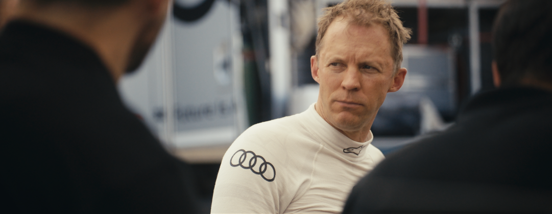 Ein junger Mann in einem weißen Rennfahreranzug mit Audi-Logo sitzt zwischen zwei anderen Personen bei einem Gespräch