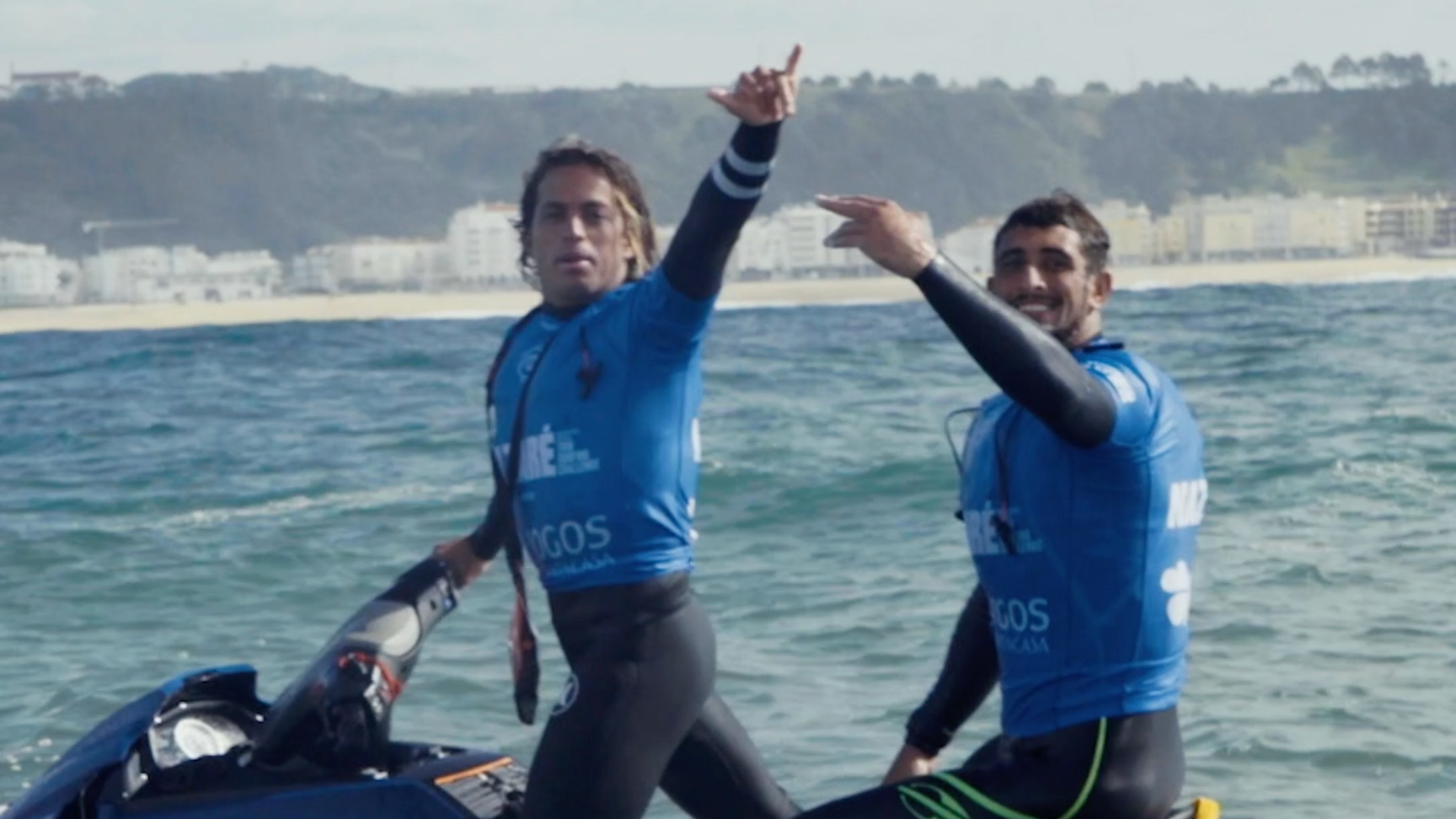 Zwei Männer auf einem Jet-Ski im Wasser, beide tragen blaue Neoprenanzüge und lachen, im Hintergrund eine Küste mit Gebäuden.