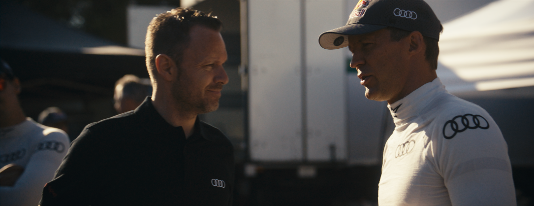Zwei Männer im Gespräch, einer trägt eine schwarze Polo mit Audi-Logo, der andere einen weißen Rennanzug und Cap, im Hintergrund sind weitere Personen und ein Truck zu sehen.
