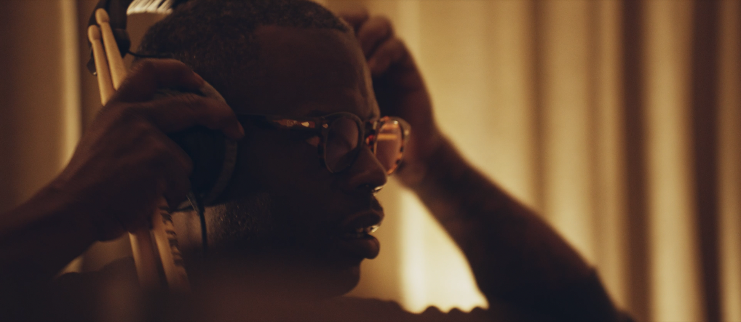 Nahaufnahme eines Mannes mit Brille, der Kopfhörer aufsetzt, bei warmem Licht in einem Raum.