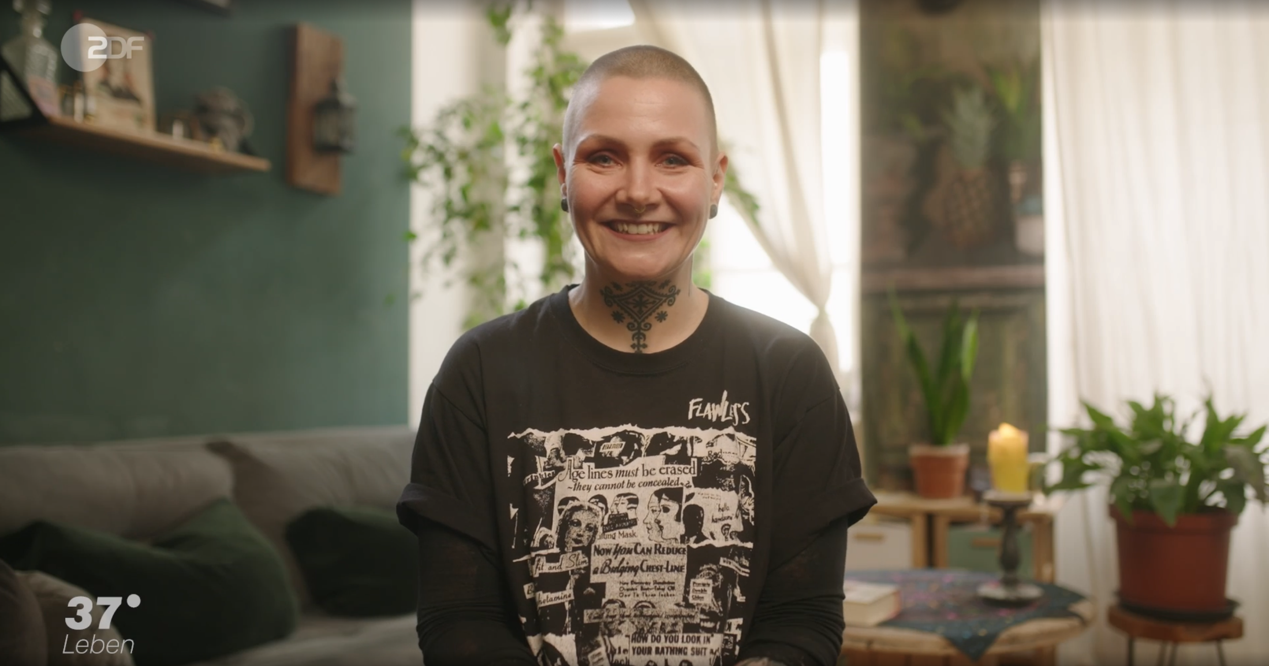 Eine lächelnde Frau mit kurzen, glatzköpfigen Haaren, schwarzen Tattoos am Hals, trägt ein schwarzes T-Shirt mit weißem Druck, sitzt in einem gemütlichen Wohnzimmer mit grünen Wänden, Pflanzen und Kerzen im Hintergrund.
